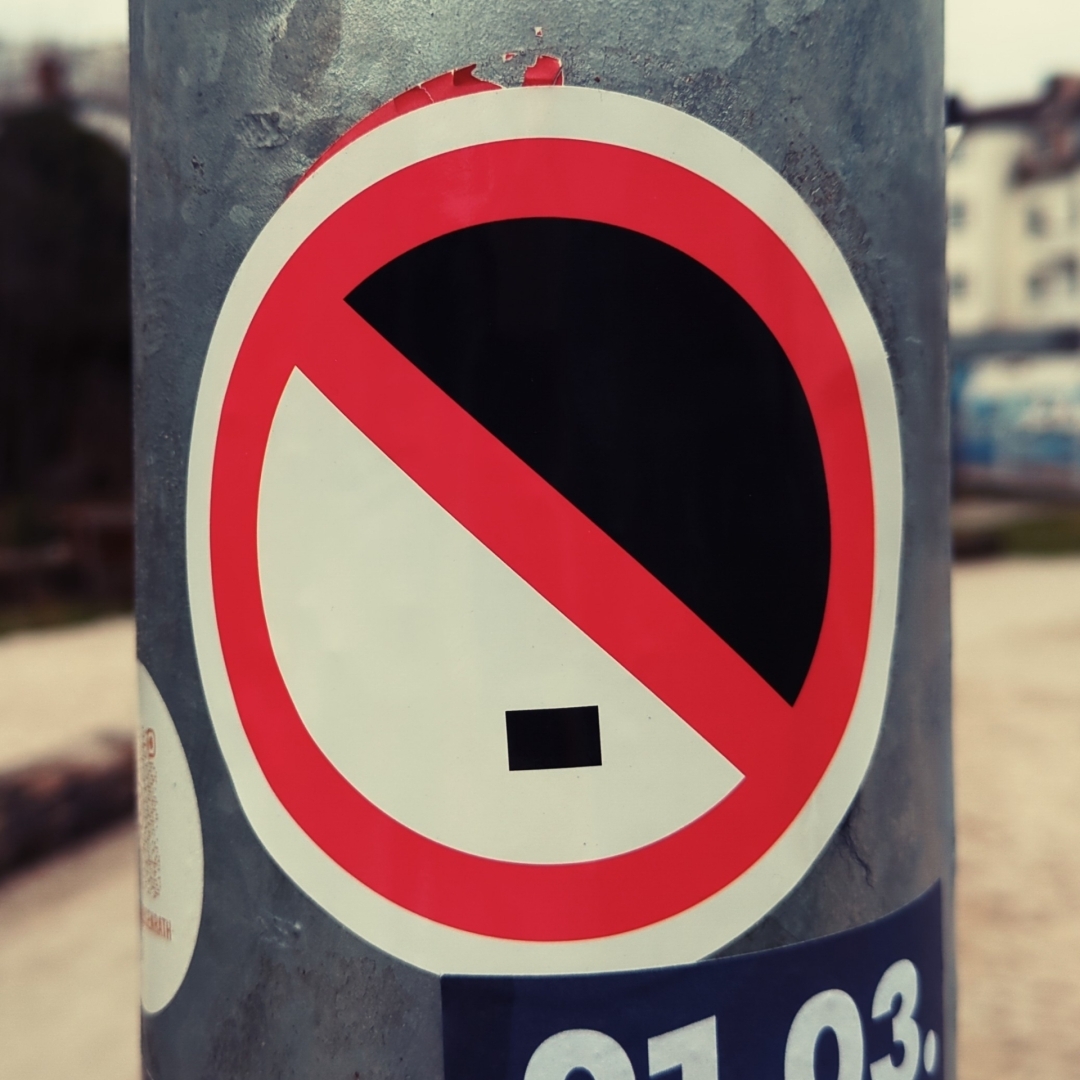 Sticker auf einem Laternenpfahl.
Das Motiv zeigt einen abstrahierten Hitlerbart und -frisur in einem Verbotsschild.